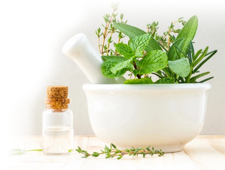 A white mortar and pestle holding fresh green herbs, with a small cork-topped glass bottle and additional herbs on the table beside it.